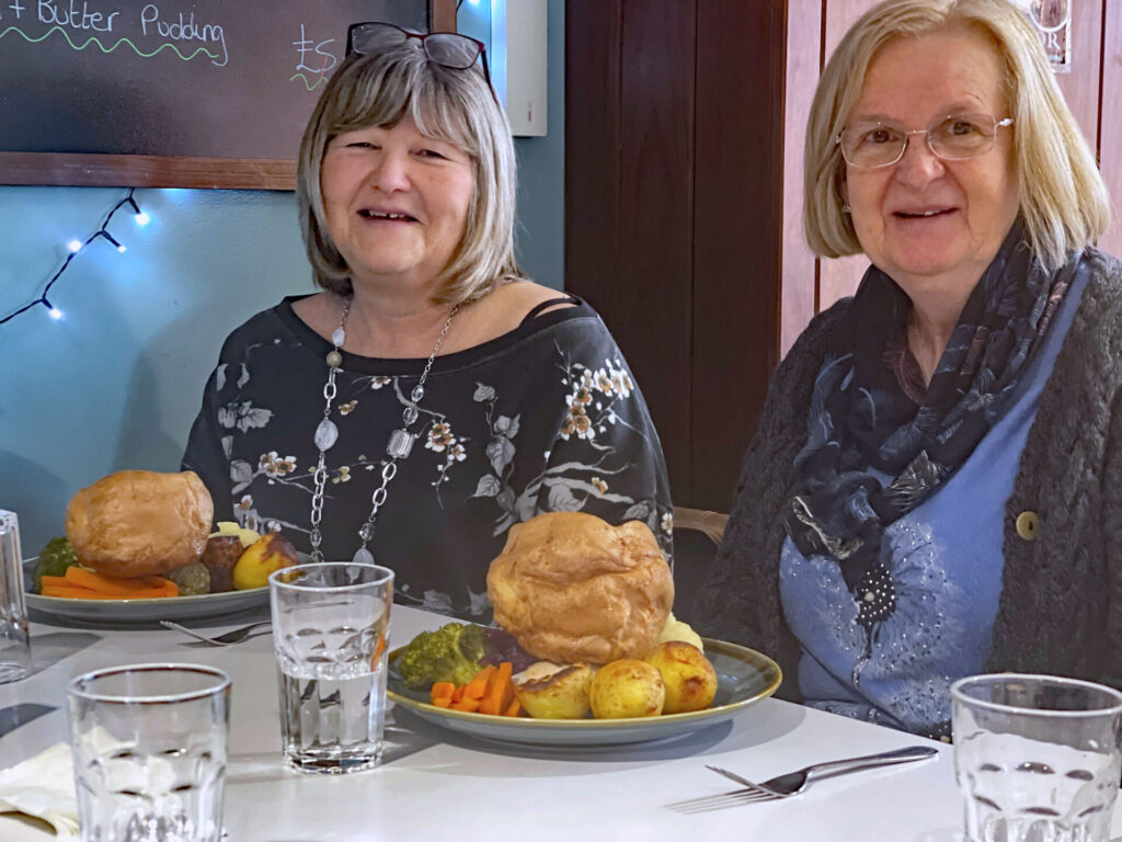 two women at a table with plates of food and glasses of water in from of them. Carrots, roast potatoes, broccoli, large Yorkshire puddings on the plates.